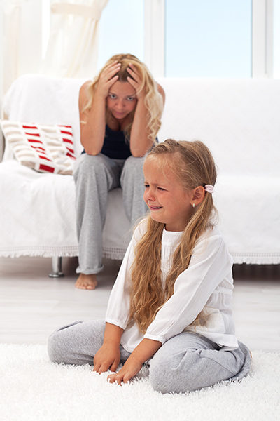 Little girl crying sitting on the floor. Mom with frustrated look sitting on couch.